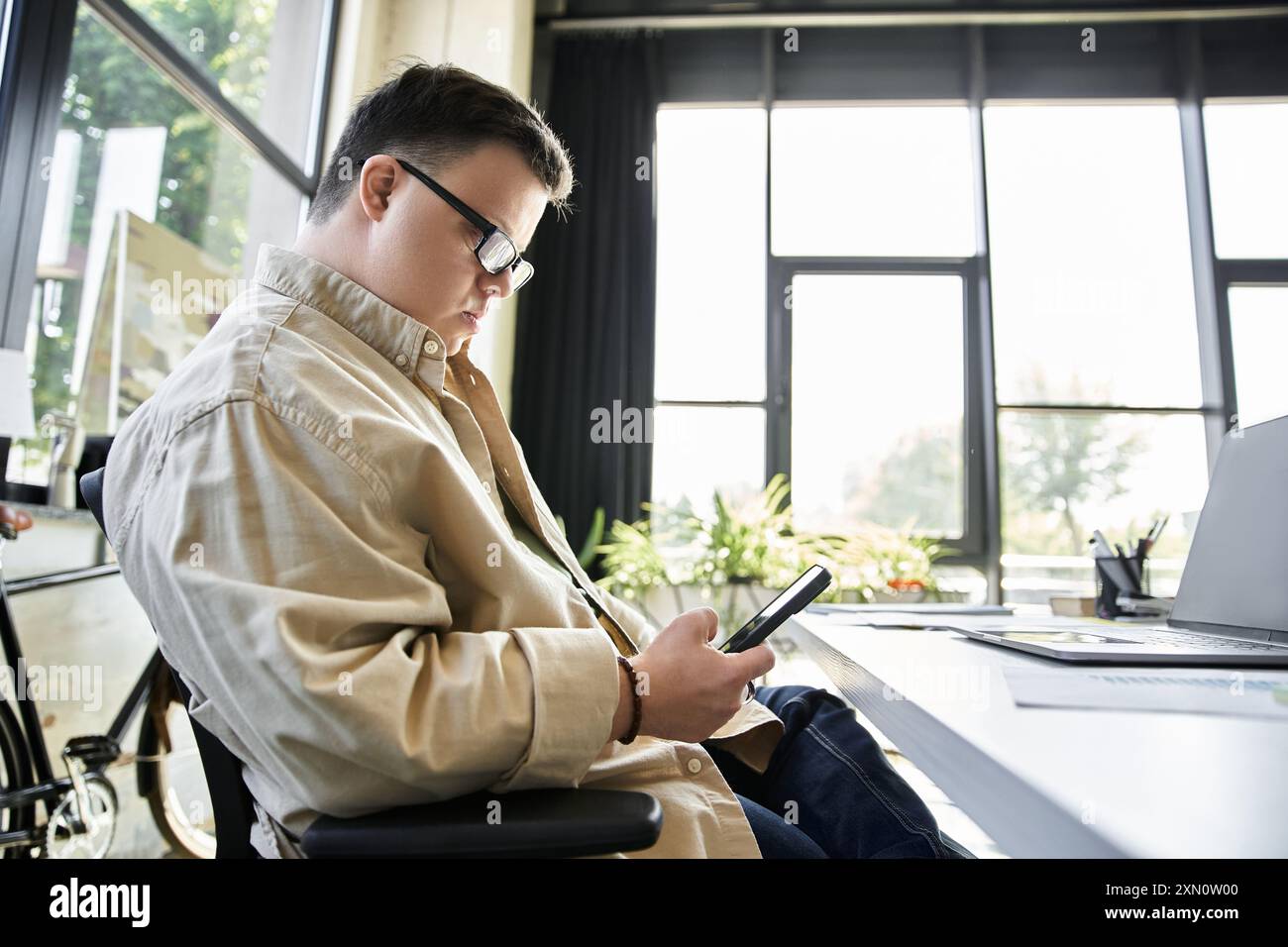 A young man with Down syndrome sits in an office chair, using his smartphone. Stock Photo