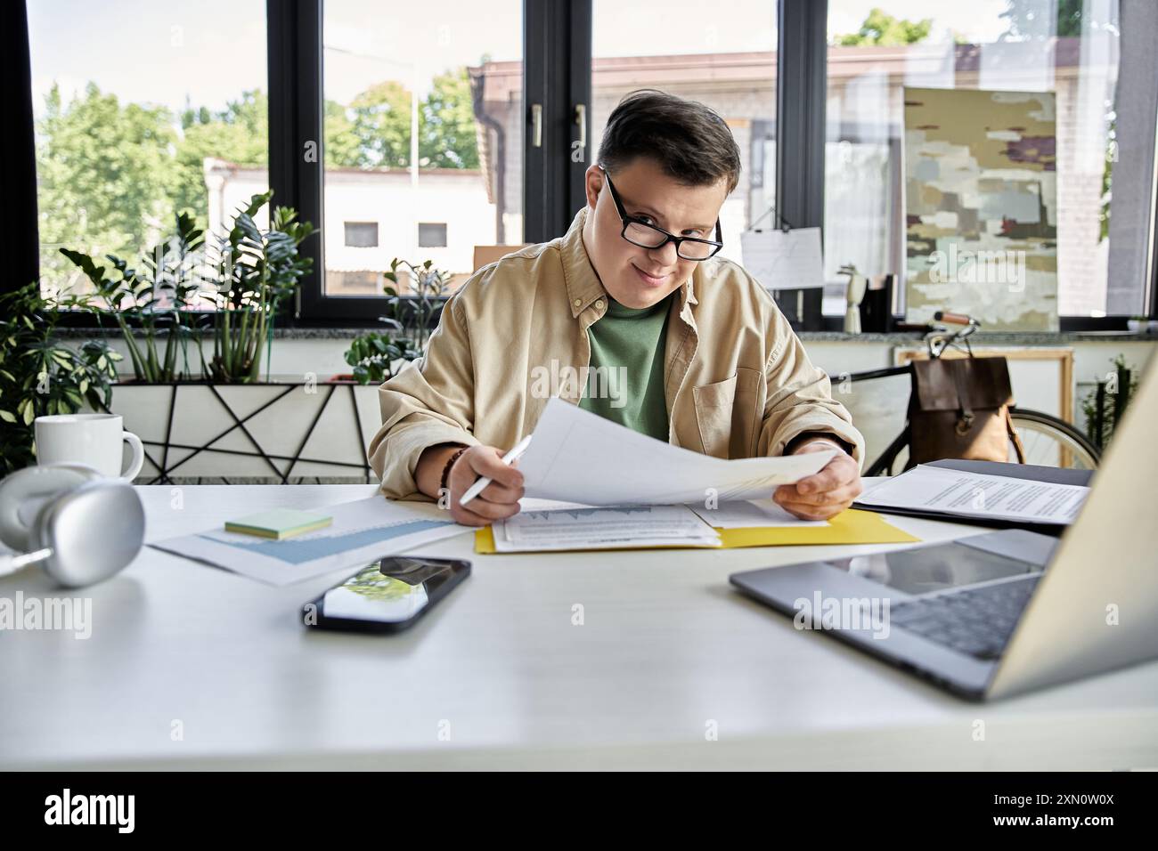 A young man with Down syndrome sits at his desk, reviewing documents while wearing glasses. Stock Photo