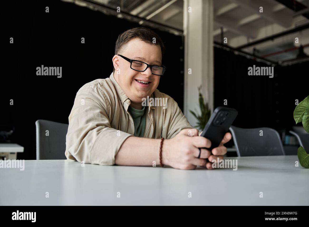 A young man with Down syndrome smiles while using his phone at a table. Stock Photo