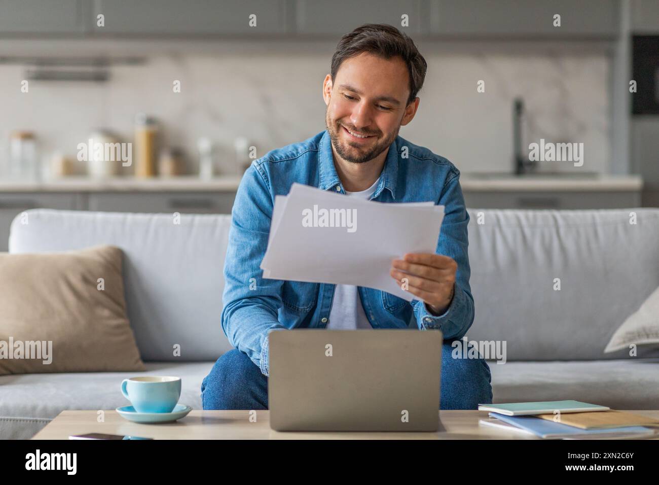 Smiling Man Reviewing Documents While Using Laptop on Sofa Stock Photo