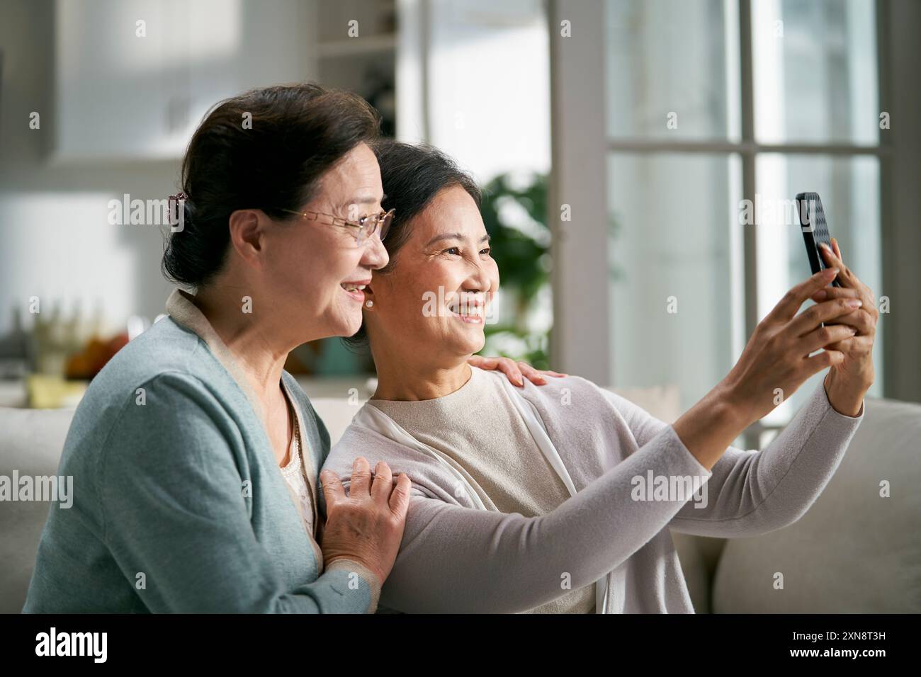 two happy senior asian women sitting on couch at home taking a selfie using cellphone happy and smiling Stock Photo