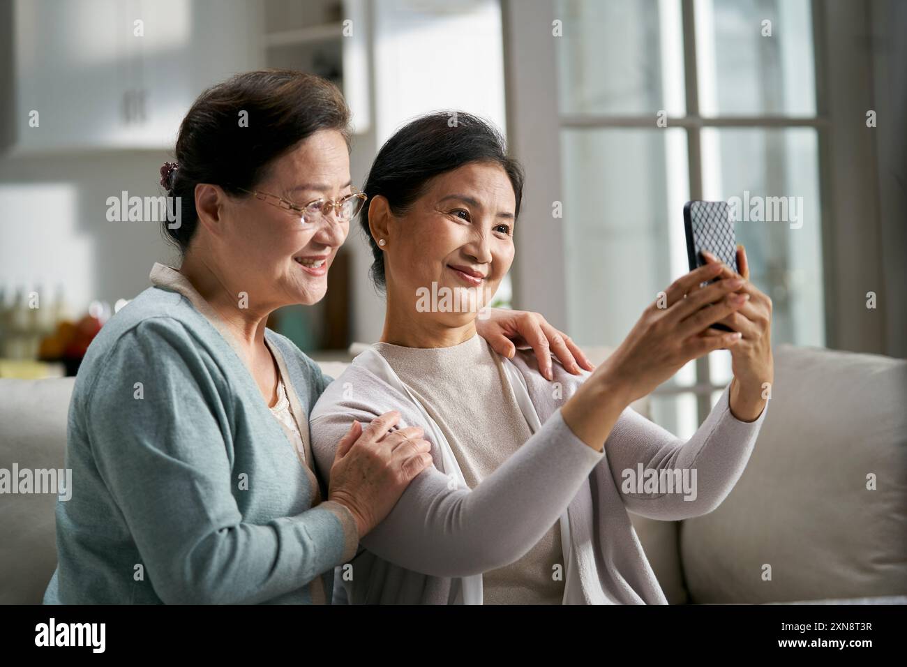 two happy senior asian women sitting on couch at home taking a selfie using cellphone happy and smiling Stock Photo