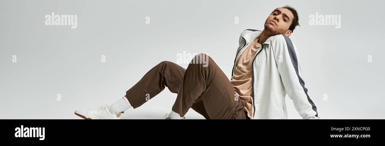 A young African American man in a stylish white jacket and brown pants poses against a grey background. Stock Photo