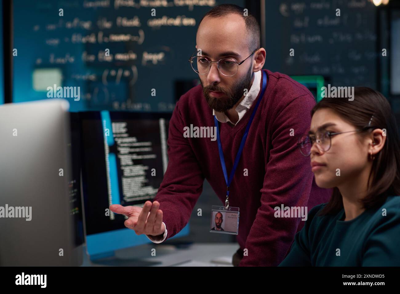 Portrait of bearded IT developer pointing at computer screen while advising young trainee in office with code lines in background Stock Photo