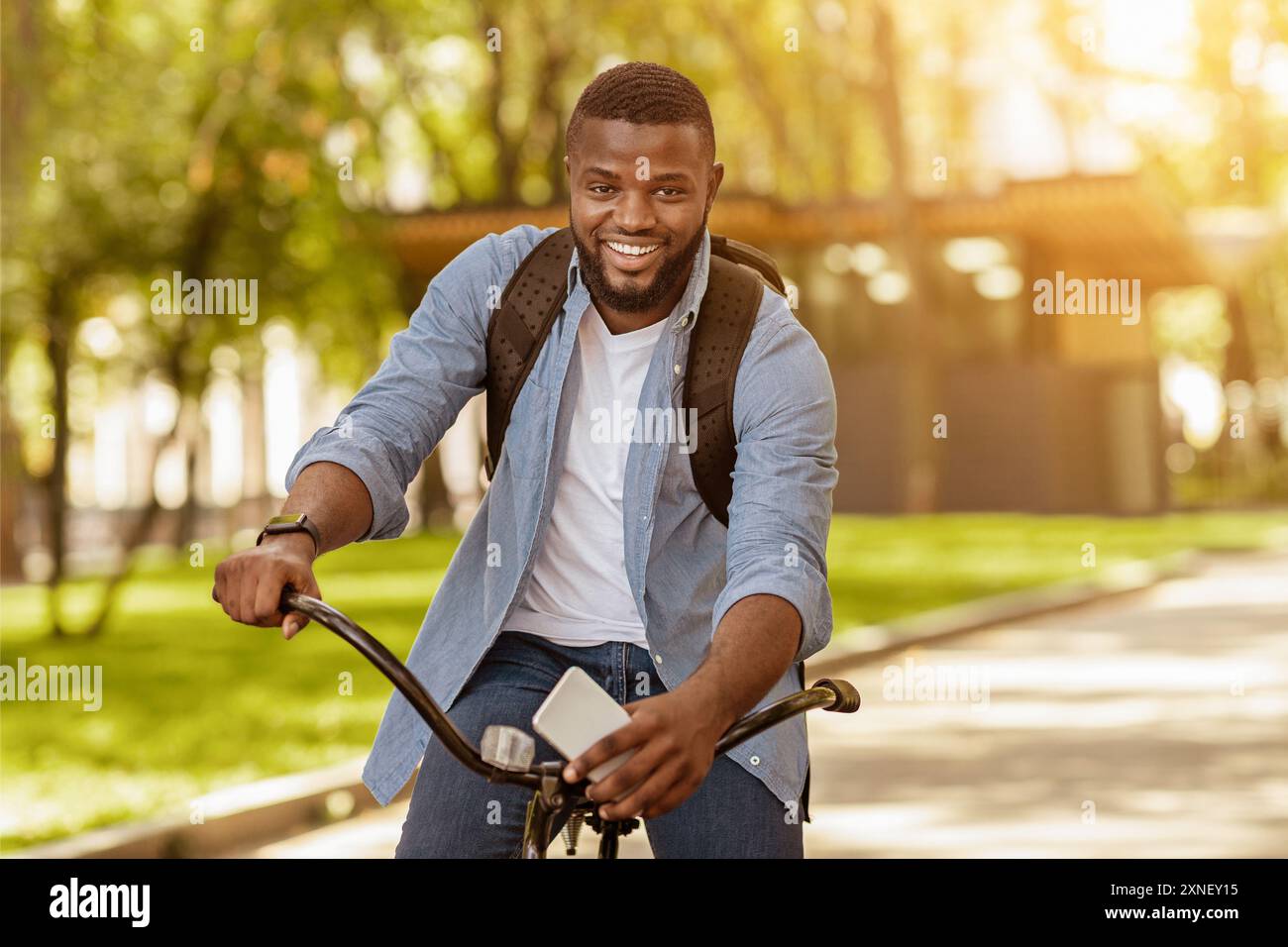 Handsome Black Man With Backpack And Smartphone Riding Bicycle, Posing Outdoors Stock Photo