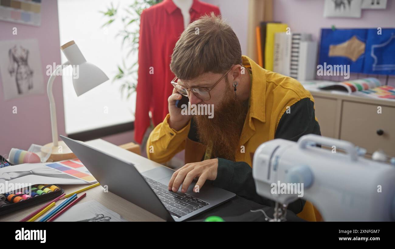 Bearded man using laptop and phone in a tailor's atelier with sewing equipment and colorful fabrics Stock Photo