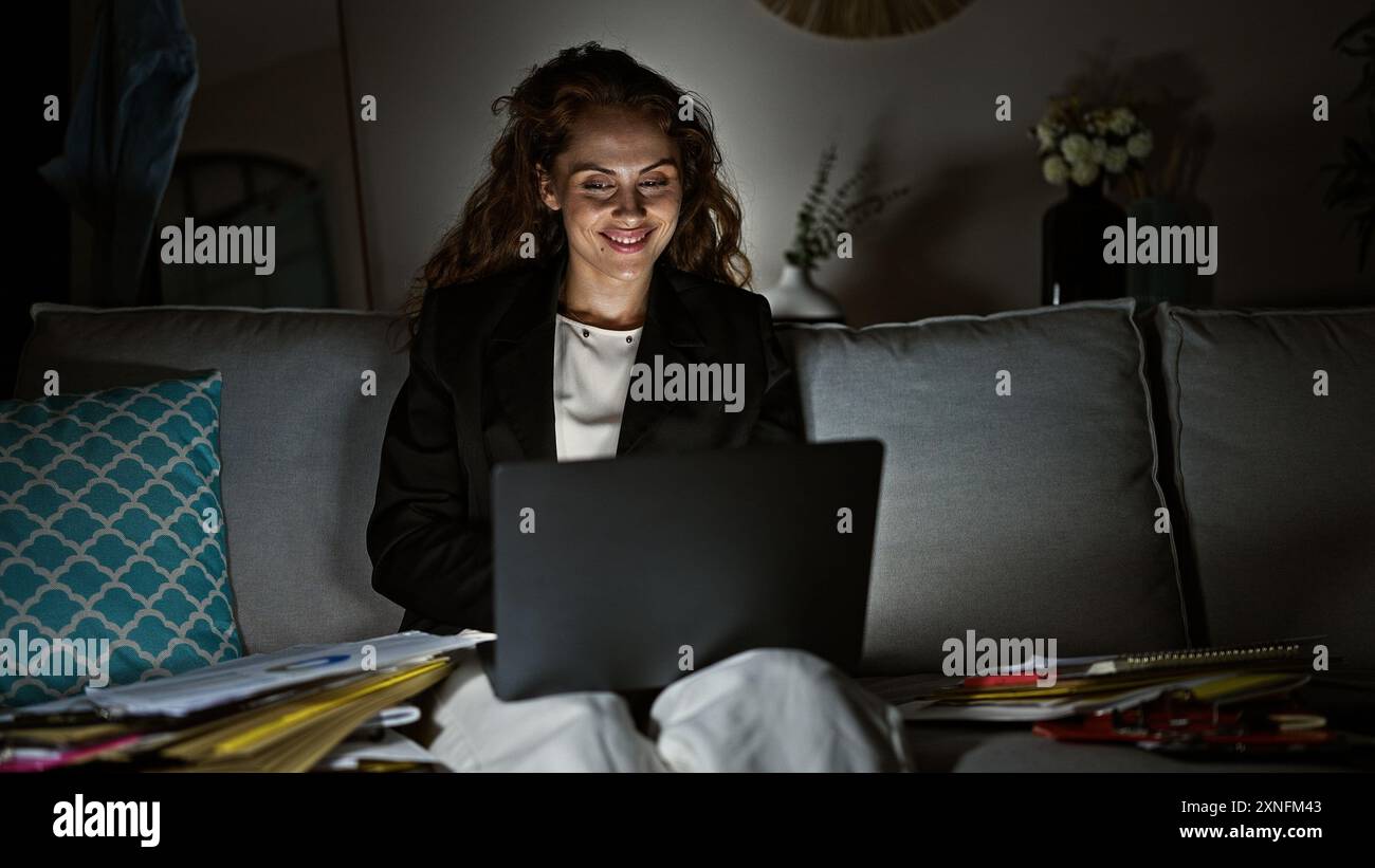 A young woman works on her laptop at night in a cozy living room ...