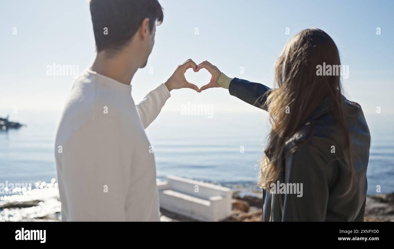 A couple forms a heart shape with their hands against a serene ocean ...