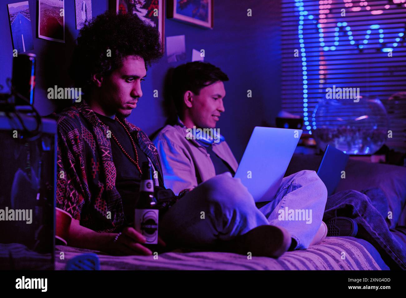 Focused roomies sitting on single bed and drinking beer while working on joint IT project Stock Photo