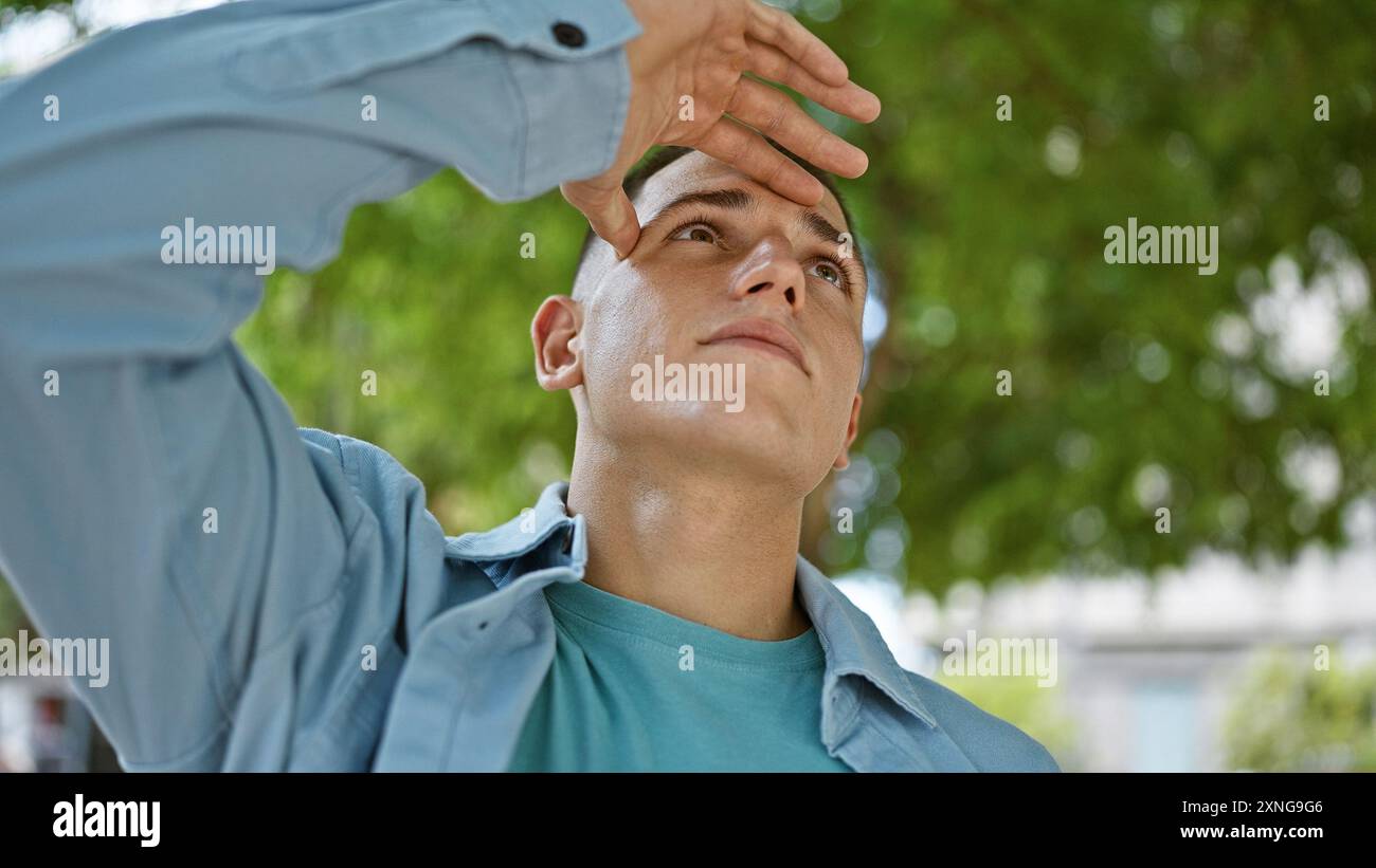 Hispanic young man shading eyes with hand, looking up in sunny park ...