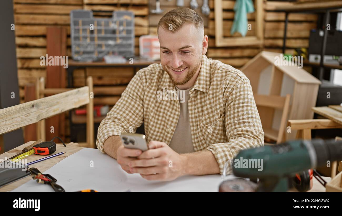 A cheerful young man using smartphone in a cozy carpentry workshop surrounded by tools. Stock Photo