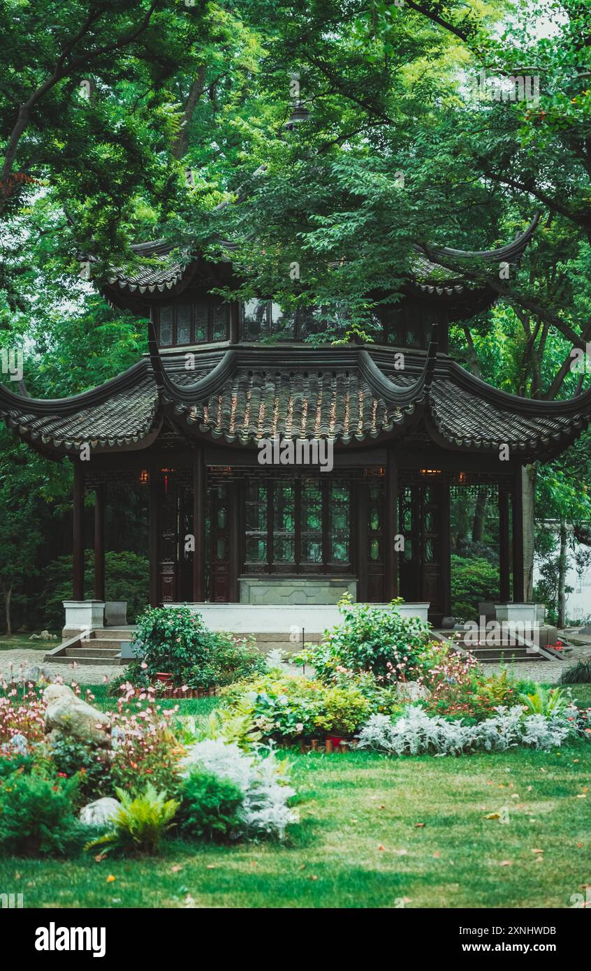 A traditional Chinese garden pavilion in Suzhou, China, with a curved roof and intricate details. Surrounding it are lush greenery, flower beds, and a Stock Photo