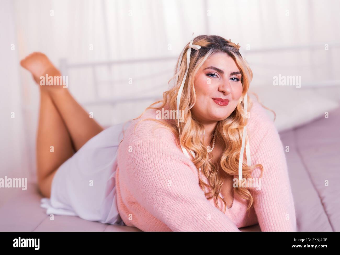Plus size woman with blond hair wearing pink sweater lying on bed and slightly smiling Stock Photo