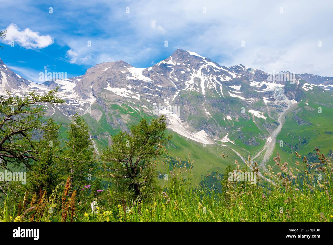 Austrian mountains in Austria with snow-capped peaks and green slopes, streams flowing down from the mountains, against a blue sky with clouds. Stock Photo