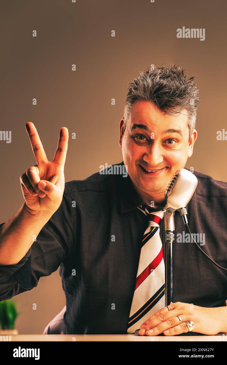 Smiling radio host confidently makes a victory sign while speaking into a vintage microphone, showcasing a positive and engaging demeanor in his workp Stock Photo
