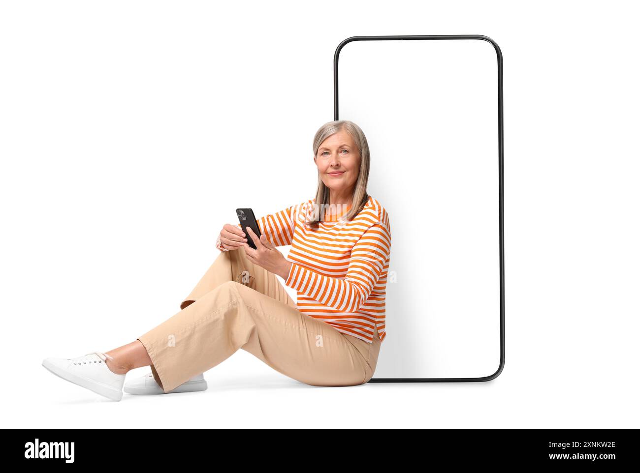 Happy mature woman holding mobile phone and sitting near big smartphone on white background Stock Photo