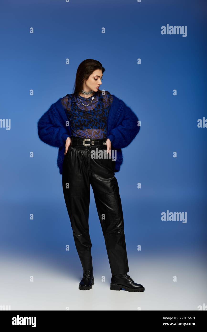 Woman in blue cardigan and leather trousers stands against blue backdrop. Stock Photo