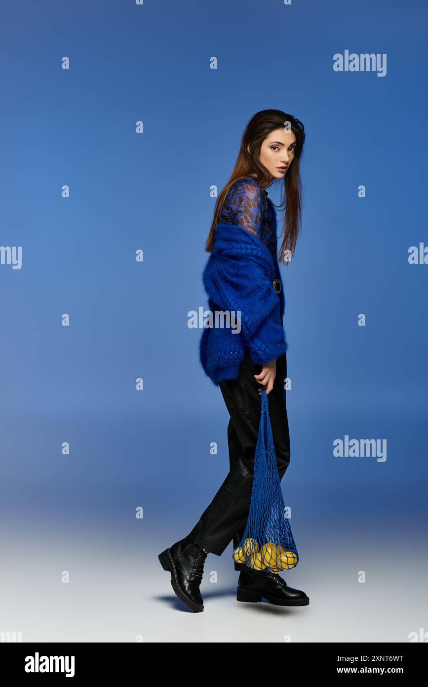 A young woman in a blue coat and black pants walks with a blue net bag filled with lemons. Stock Photo