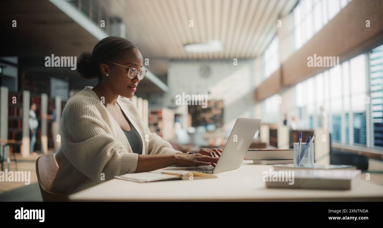 Dedicated African American Female Student Engaged in Online Learning at a Modern Library. Young Woman Using Laptop for Research, Surrounded by Books and Academic Environment. Stock Photo