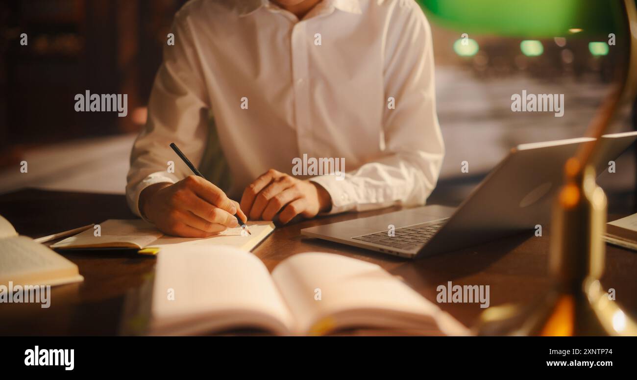 Dedicated Male Student Engaged in Academic Research With Books and Laptop in a Cozy Library Setting. Young Man Writing Notes, Focused on E-Learning and Preparing for Exams. Stock Photo