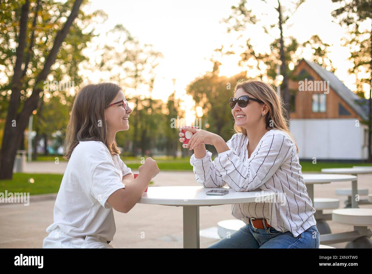 Cheerful adult woman and teenager smiling and enjoying fresh coffee while sitting at table in park cafe in evening Stock Photo
