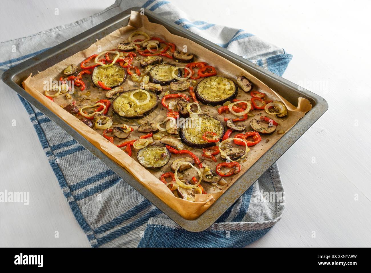 Oven vegetables with herbs and spices on a baking tray placed on a blue kitchen towel and a white table, Mediterranean vegetarian meal, copy space Stock Photo