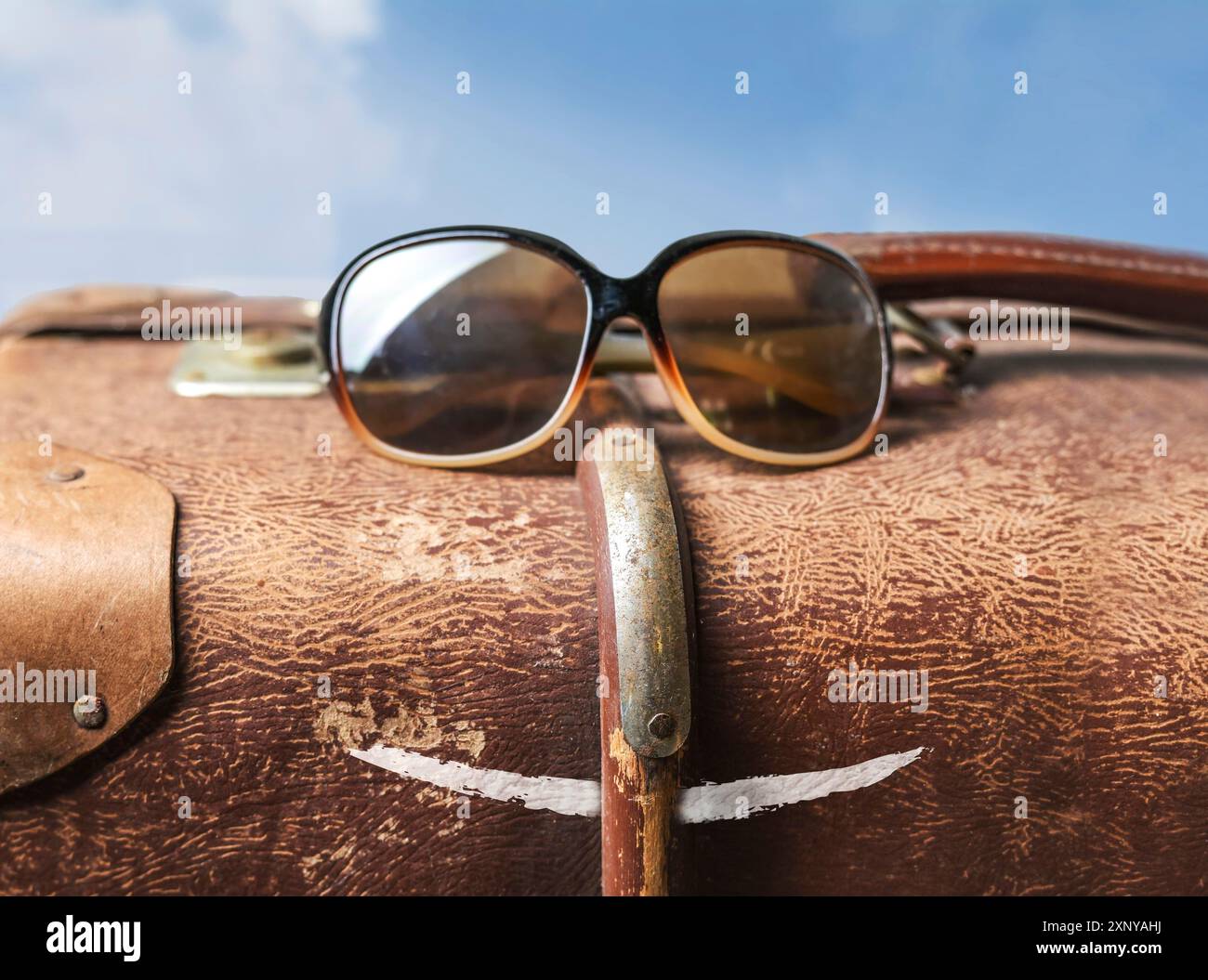 Funny travel face from an old brown suitcase with sunglasses and a painted mouth against a blue sky with clouds, copy space, selected focus, narrow Stock Photo