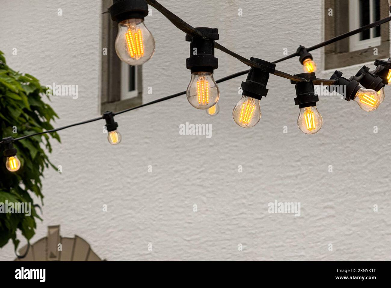 A strand of glowing, decorative Edison light bulbs illuminates a quaint European courtyard at dusk. Stock Photo