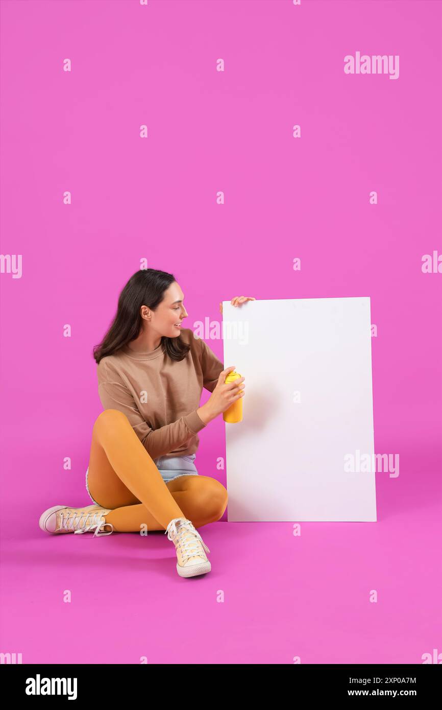 Young woman with spray paint can and blank paper sitting on purple background Stock Photo