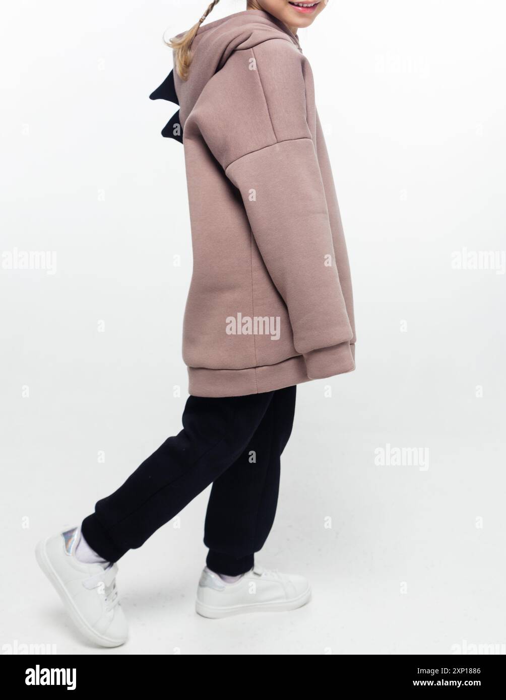 A child walking while wearing a beige hoodie with a black dinosaur tail detail and black pants, smiling against a white background. Stock Photo