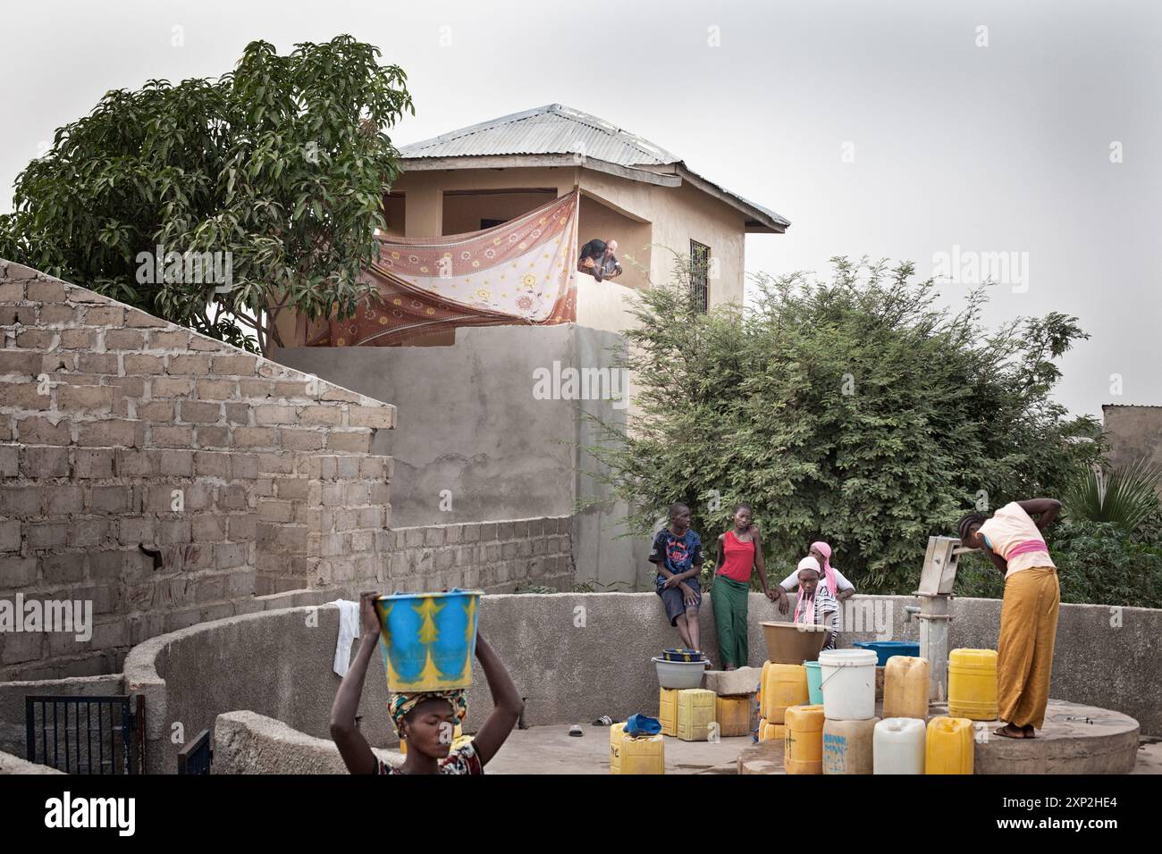 Women in a Gambian village fetching water from a well while children play nearby and another woman watches from a balcony. Stock Photo