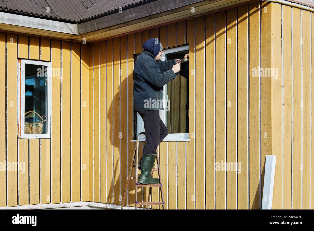 Roofing contractor does exterior trim of window opening while standing on steel step ladder, in front of facade of wooden country house on an autumn a Stock Photo