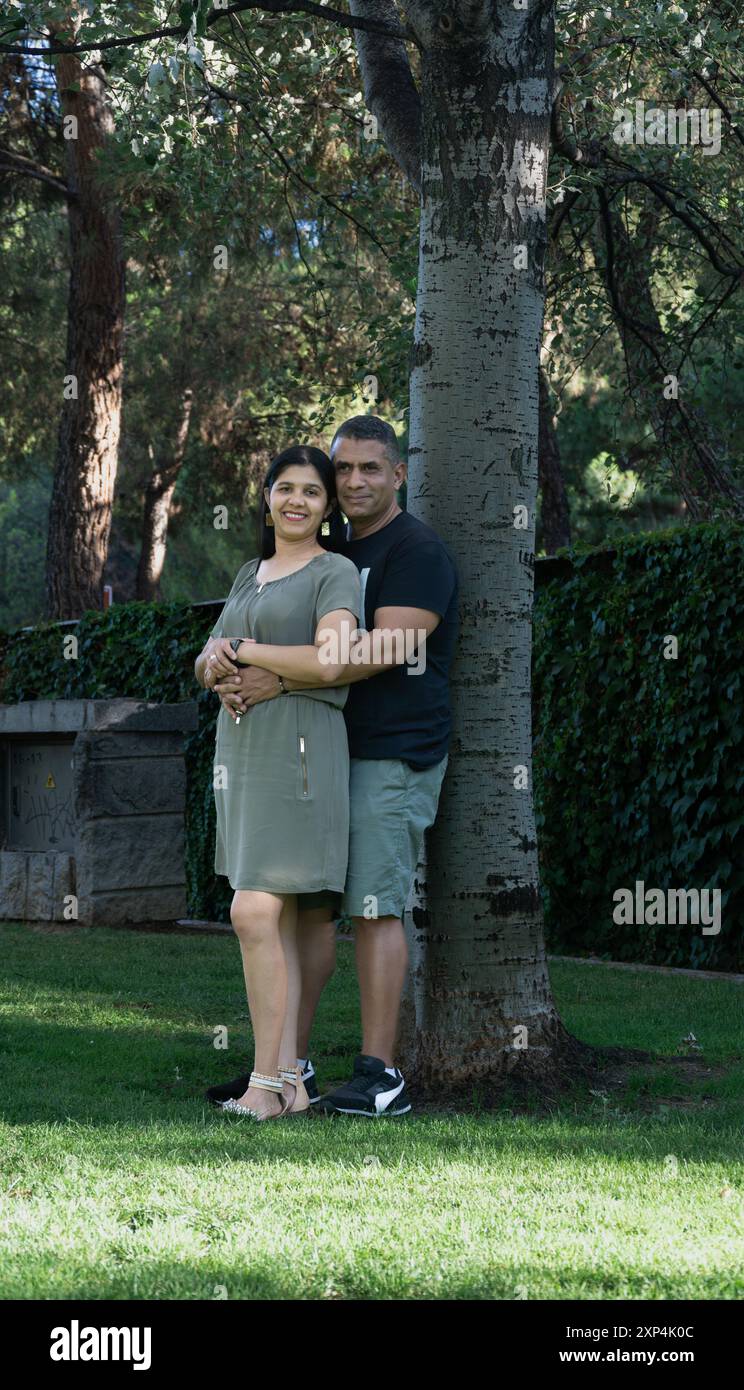 A man and woman are standing next to a tree in a park. They are smiling ...