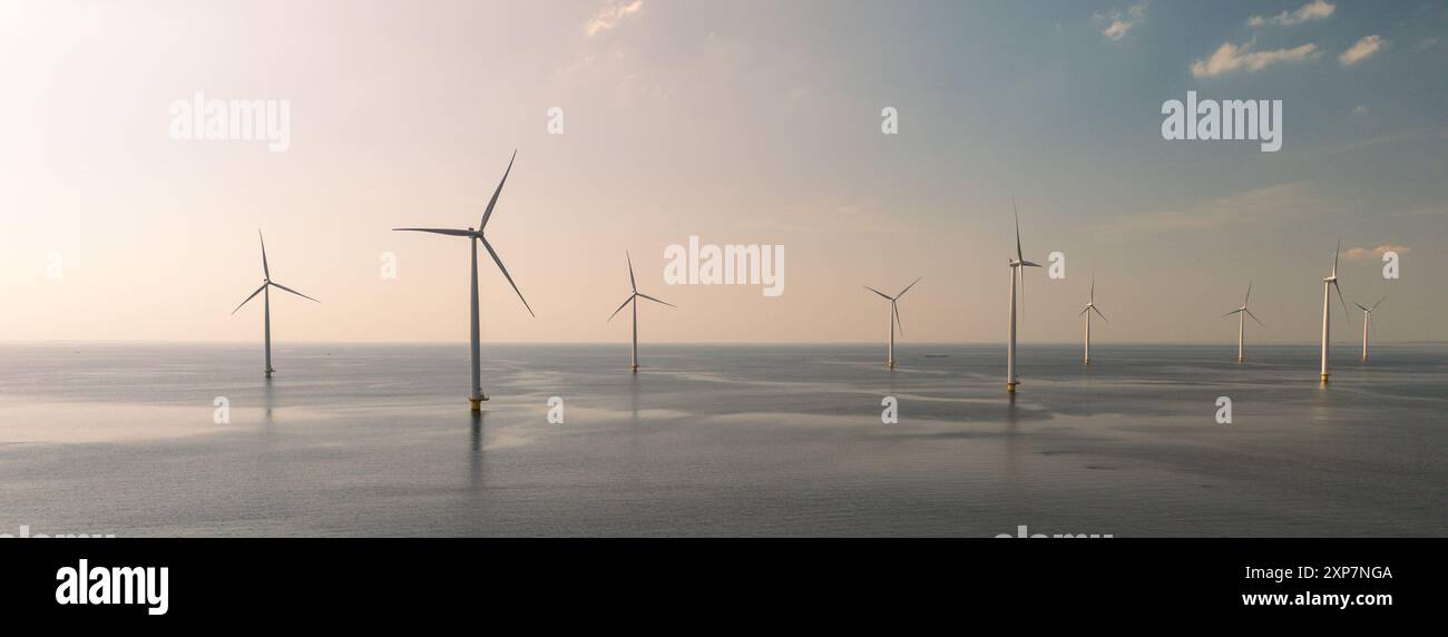 Towering windmills gracefully spin against a tranquil backdrop, reflecting the soft hues of dusk over the water. windmill park in the Netherlands, green energy , energy transition Stock Photo