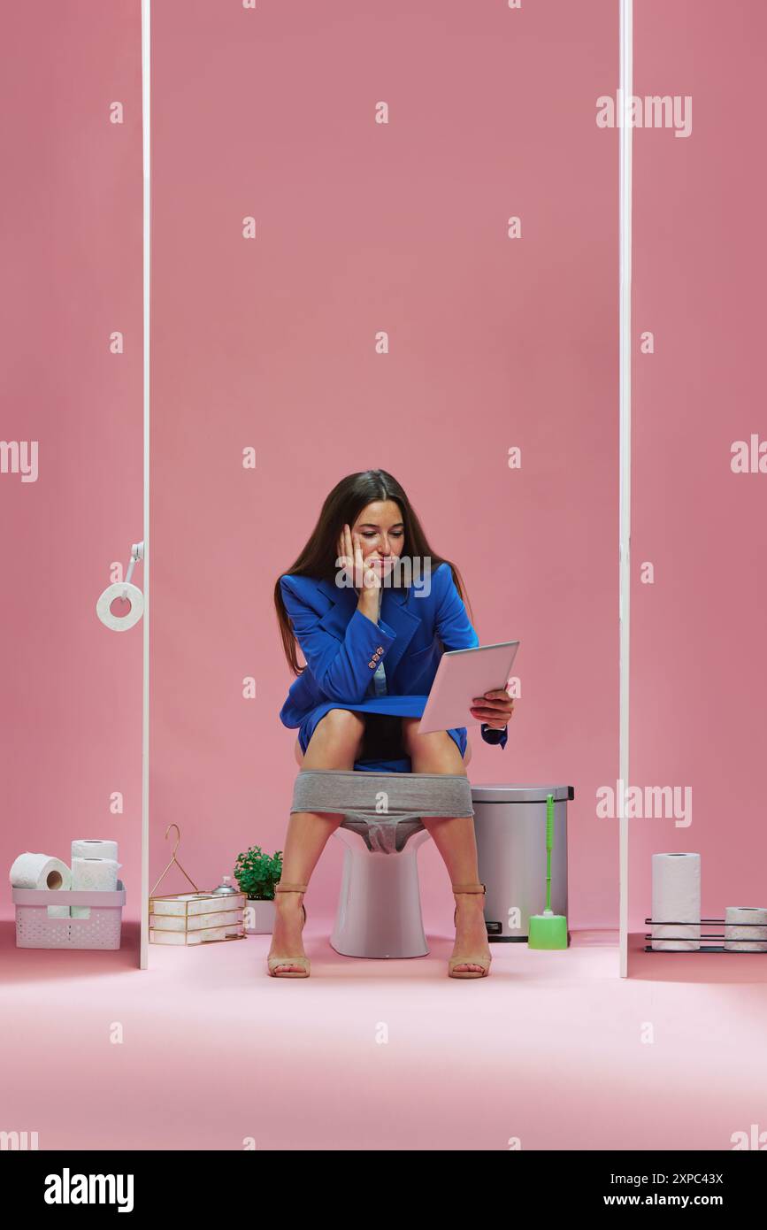 Business woman in blue suit sitting on toilet and watching on tablet with bored face, waiting for soccer game against pink background Stock Photo