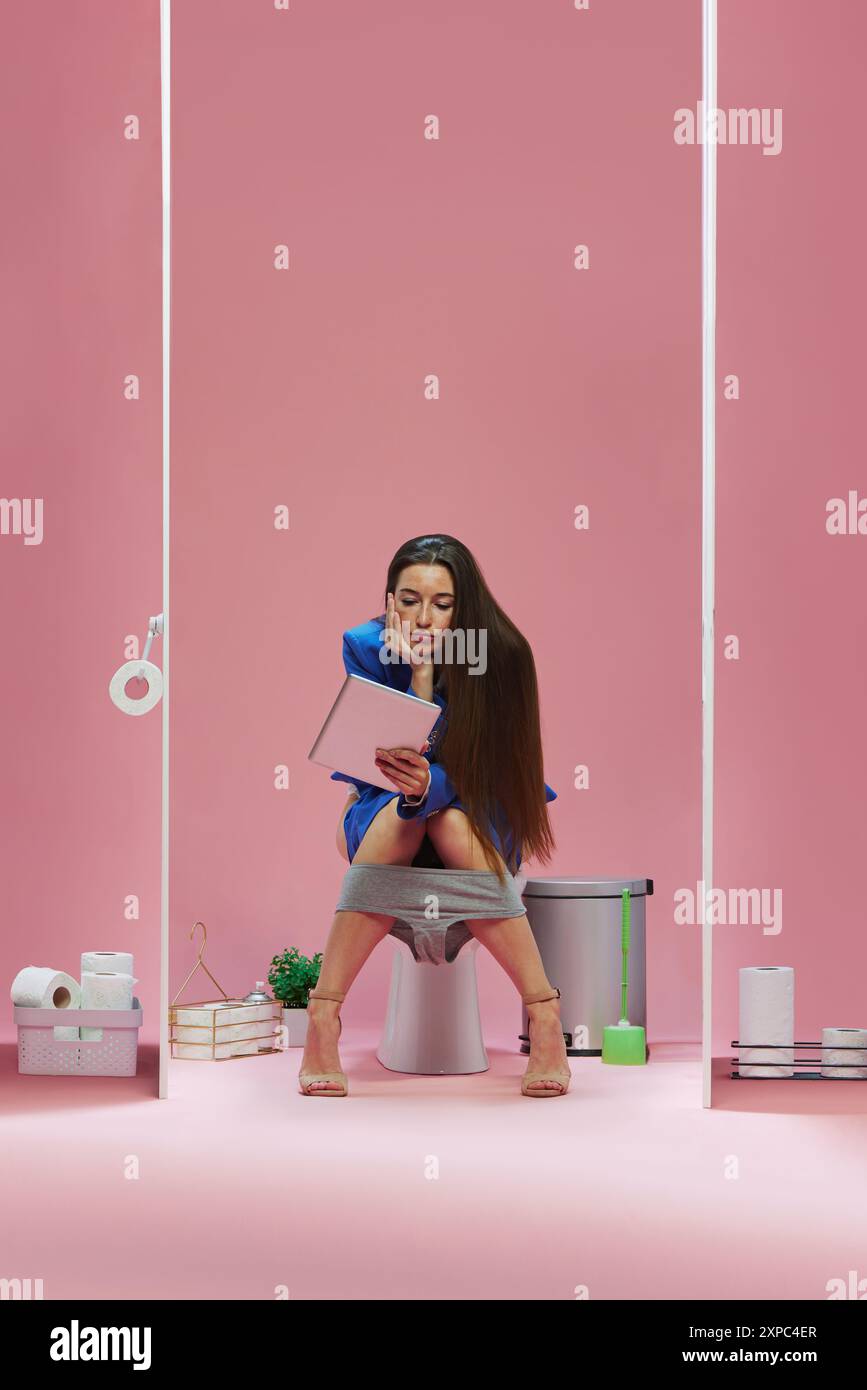 Business woman in blue suit sitting on toilet with bored face and looking in tablet against pink background Stock Photo