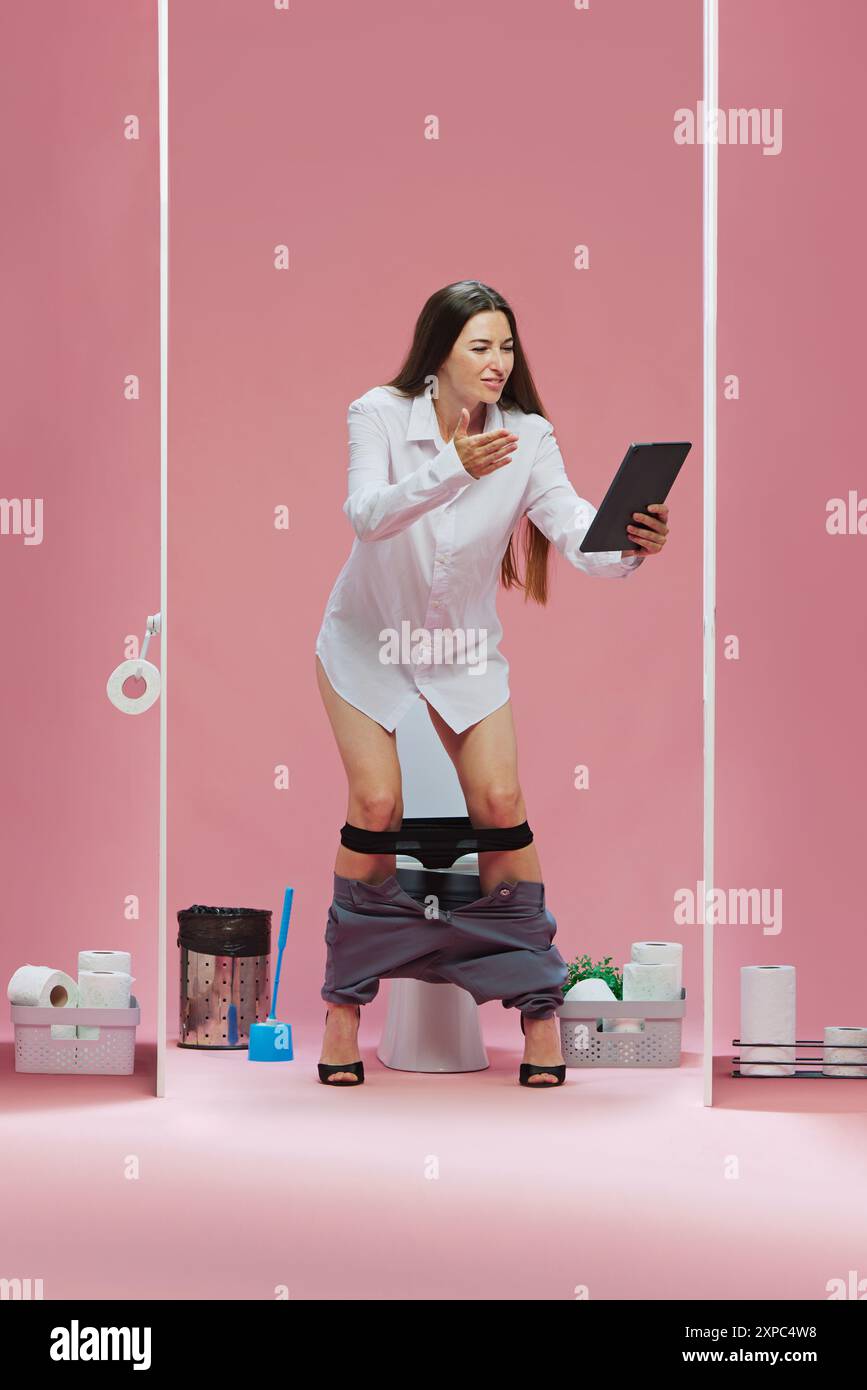 Missing goal. Young woman in white shirt standing in toilet with tablet, watching online game translation with emotional face against pink background Stock Photo