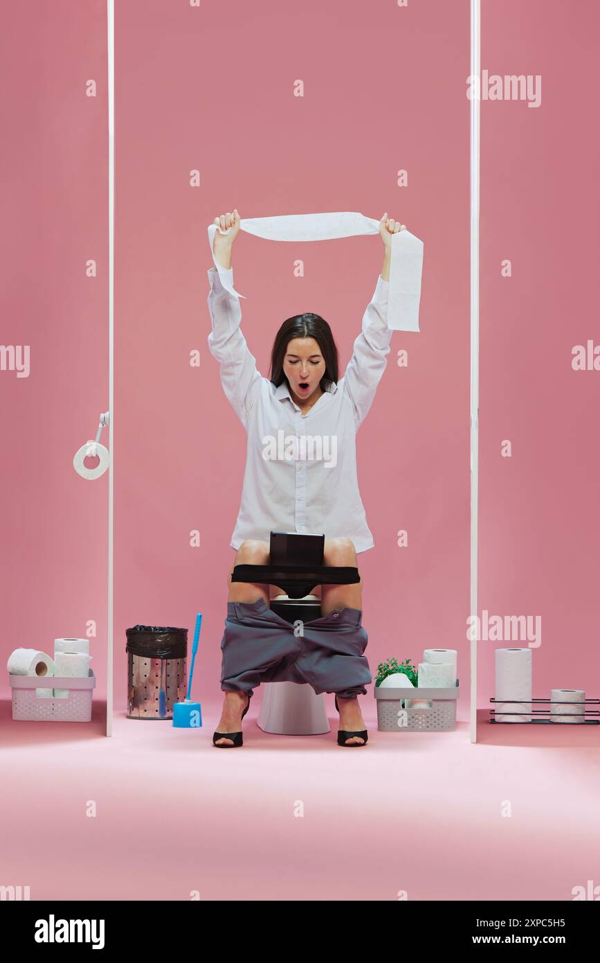 Emotional business woman sitting on toilet, raising toilet paper upwards and watching soccer game event online via laptop against pink background Stock Photo