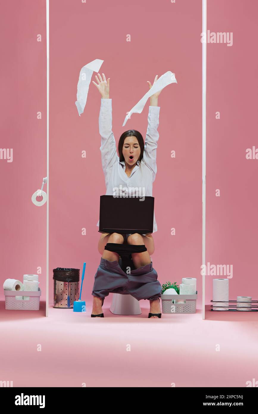 Emotional business woman sitting on toilet, raising toilet paper upwards and watching soccer game event online via laptop against pink background Stock Photo