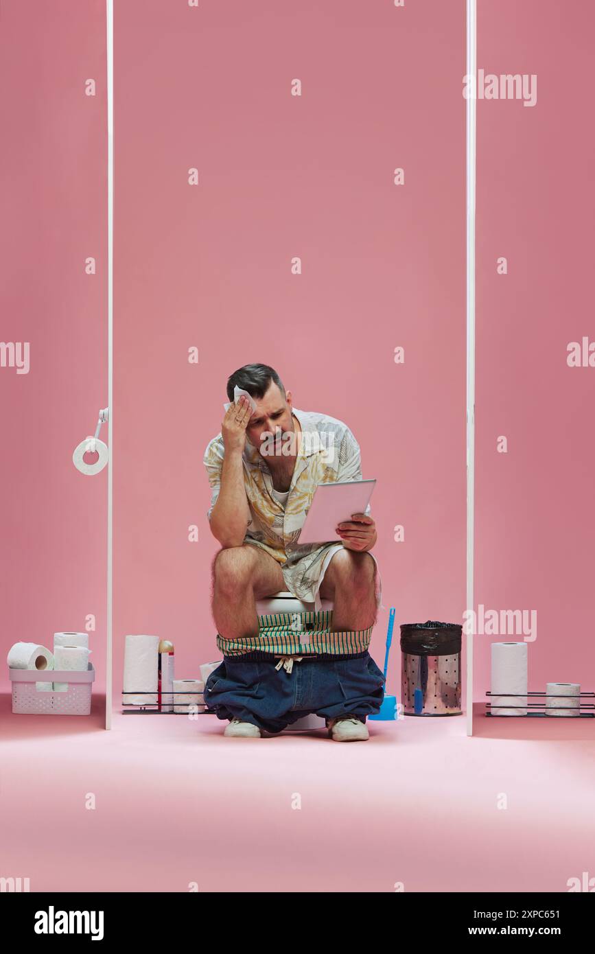 Tense game moment. Young man sitting in toilet, wiping forehead with paper and looking on tablet with sad face against pink background Stock Photo