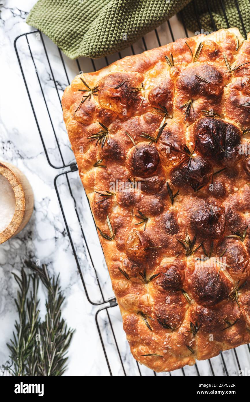 A deliciously and crispy focaccia bread, topped with sprigs of rosemary and salt flakes, cooling on a wire rack. Stock Photo