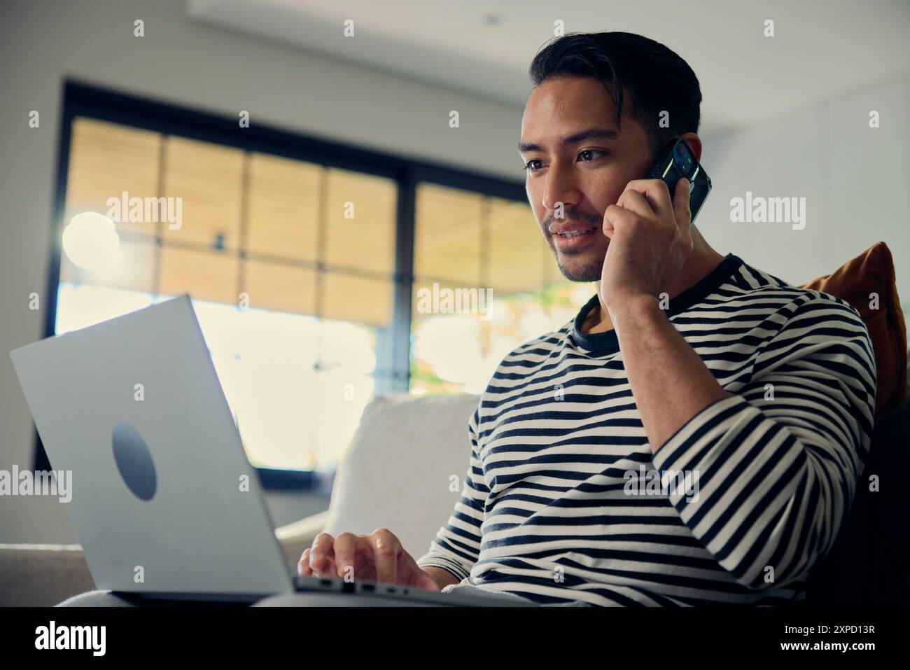 Man on couch using laptop, talking on cell phone, gesturing with arm Stock Photo