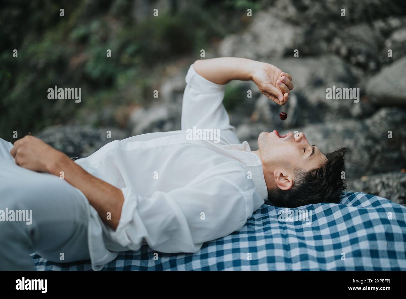 Young man lying on a picnic blanket eating cherries outdoors in nature Stock Photo