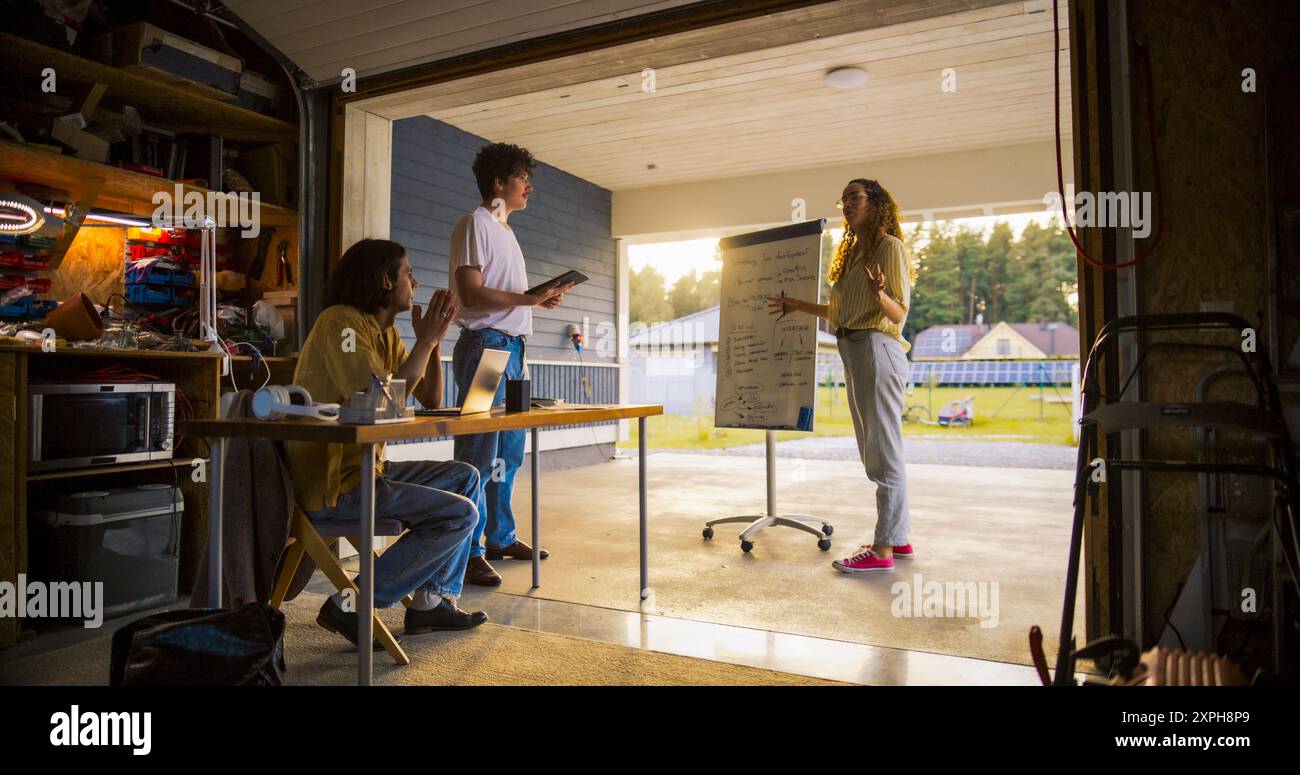 Team Of Young Entrepreneurs Having A Meeting In Home Garage. Hispanic Female Manager Using Whiteboard To Outline Business Plan For e-Commerce Startup. Two Male Colleagues Using Laptop And Tablet. Stock Photo