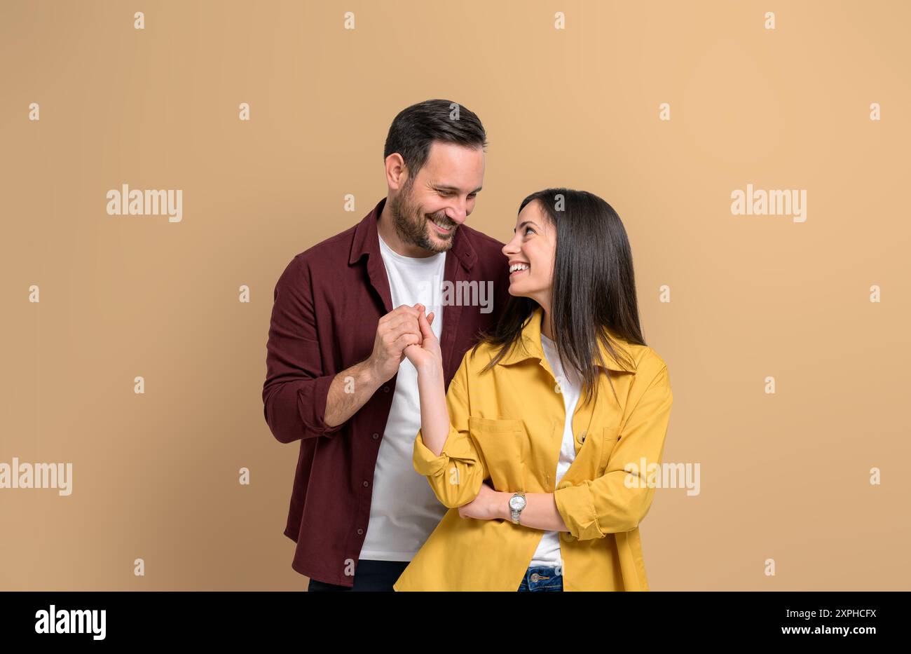 Romantic young man and woman holding hands and smiling while standing over beige background. Attractive couple dressed in casuals looking at each othe Stock Photo