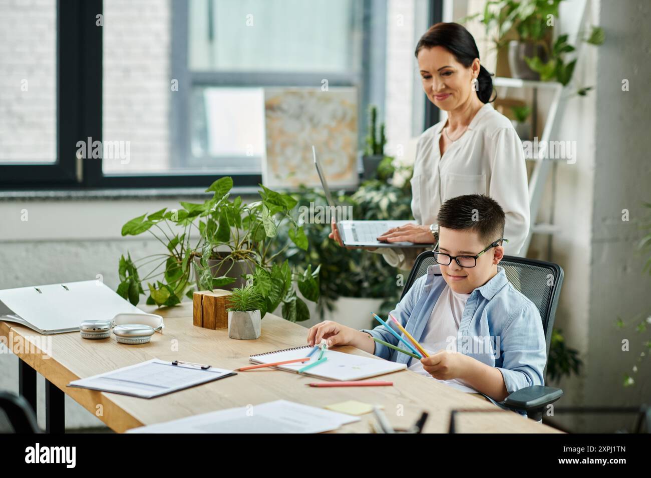 A mother works on her laptop while her son, who has Down syndrome, draws at the table in a home office setting. Stock Photo