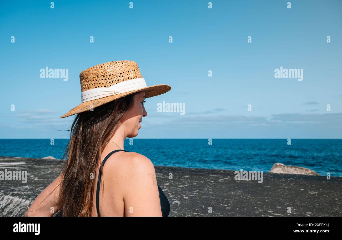 A woman wearing a straw hat stands on a beach looking out at the ocean ...