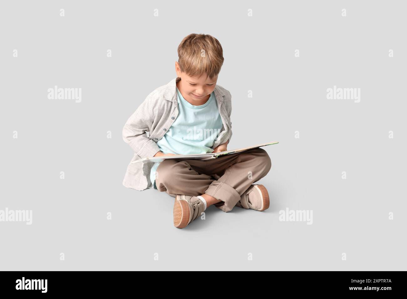 Cute little boy sitting on floor and reading book on grey background ...