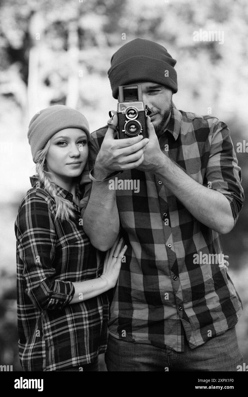 Bald guy with a beard and a blonde girl in bright hats on the background of the river are taking pictures with an old camera Stock Photo