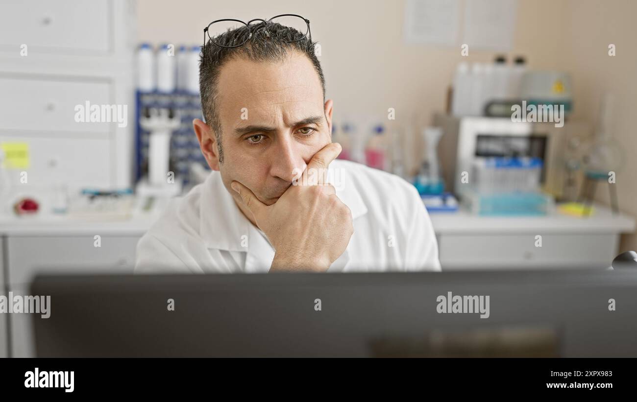 A thoughtful man in a white lab coat ponders at a computer in a medical ...